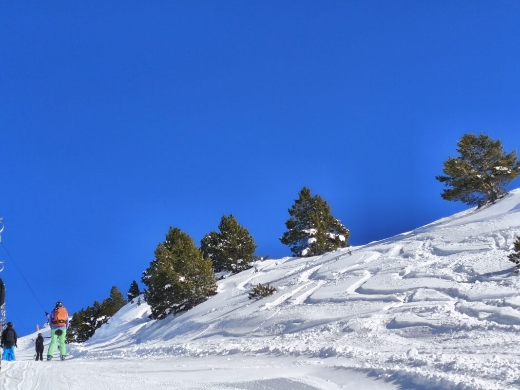 Esquiadores en la percha al lado de una pista con trazas de nieve polvo y sol en Baqueira.
