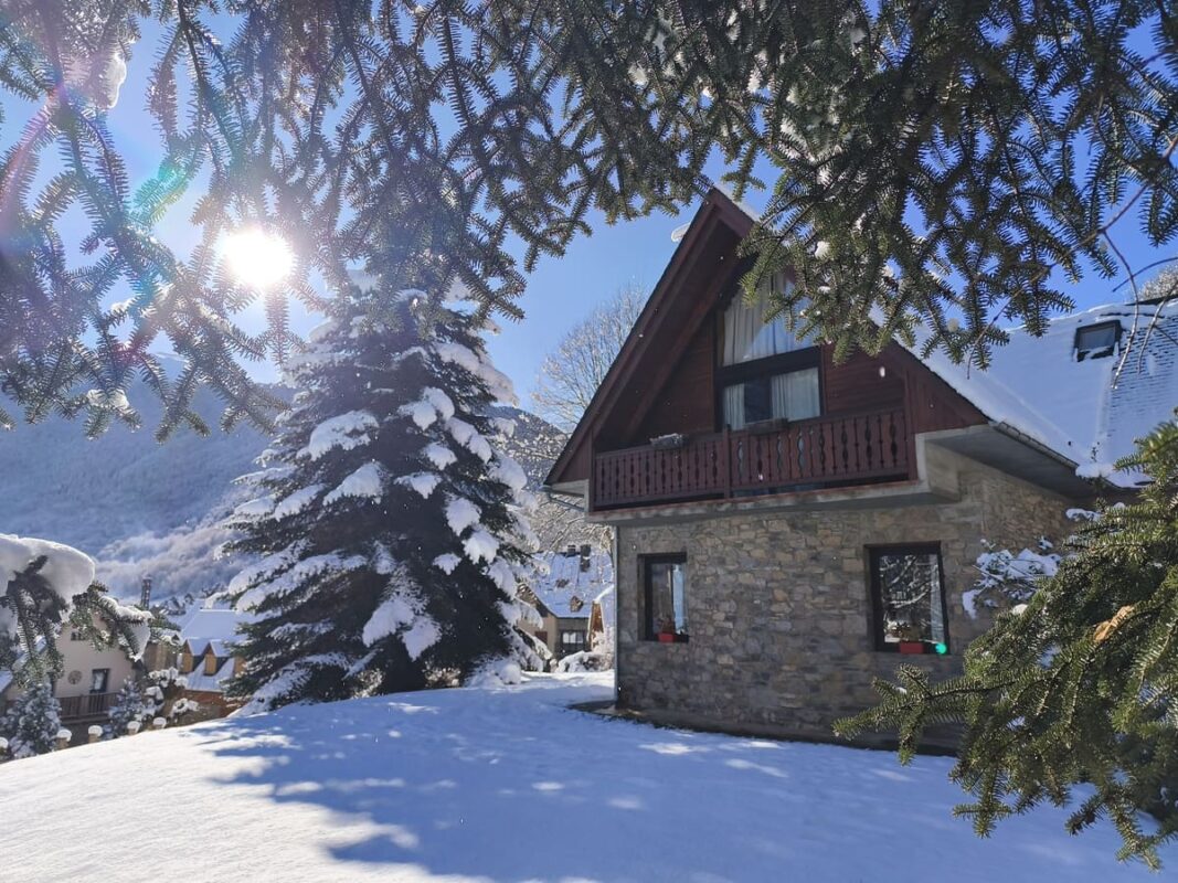 Vue romantique de la Casa Lola Pirene à travers les branches de sapins enneigés sous le soleil.